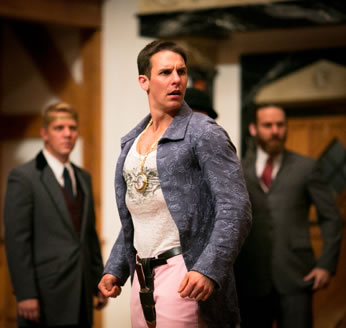 Gaveston angrily stands looking over his left shoulders, hands semi-clenched at his side, his muscular chest straining the threads of his tight T-shirt, the two lords in he background in formal three-piece gray suits, Edmun wearing a gold head band and a dark tie, Pembroke in a red tie.